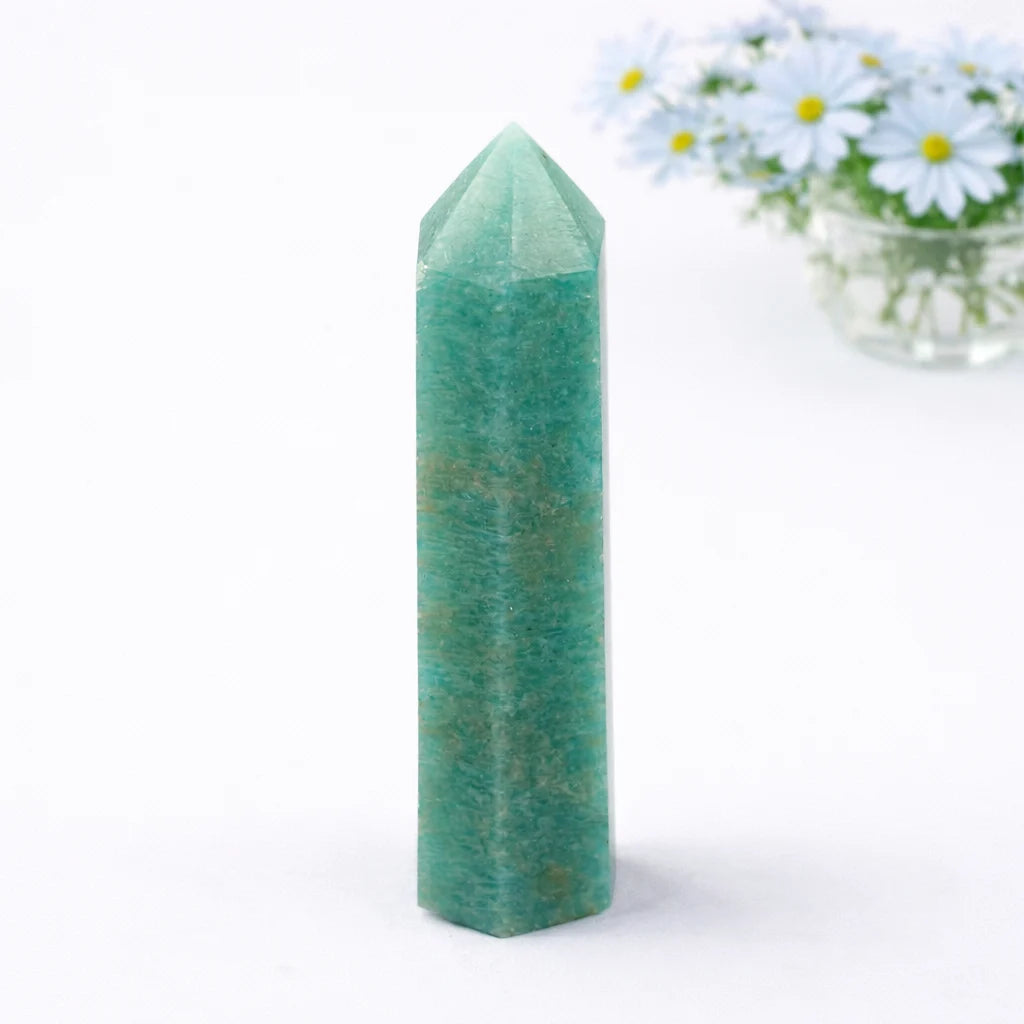 Natural amazonite polished crystal point with green-blue tones on clean white surface with subtle shadow and floral background